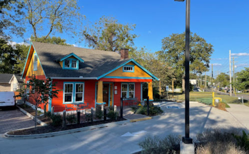 Colorful house with orange and blue accents, surrounded by trees and a parking area, under a clear blue sky.
