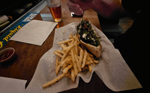 A plate with a sandwich filled with greens and tomatoes, accompanied by a side of seasoned French fries and a drink.