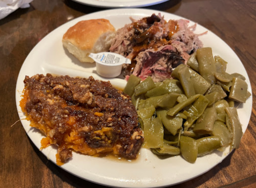 A plate of food featuring pulled pork, green beans, sweet potato casserole, a biscuit, and a sauce packet.