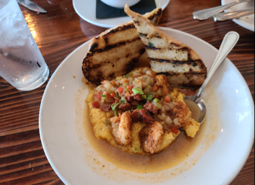 A plate of shrimp and grits topped with diced vegetables, served with two slices of grilled bread and a glass of water.
