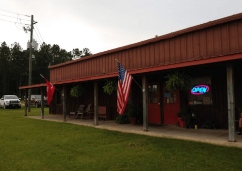 A rustic building with an "OPEN" sign, American and state flags, and potted plants along the porch.