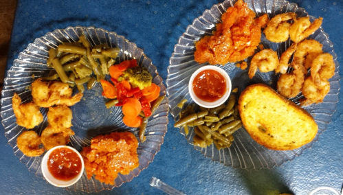 Two plates of fried shrimp and chicken, served with green beans, mixed vegetables, and dipping sauce on a blue table.