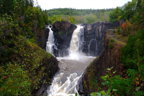 A scenic view of a waterfall cascading between rocky cliffs, surrounded by lush greenery and a cloudy sky.