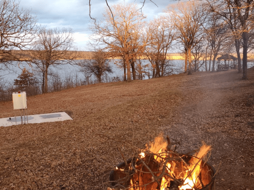 A cozy campfire burns in the foreground, with a serene lake and bare trees in the background under a cloudy sky.