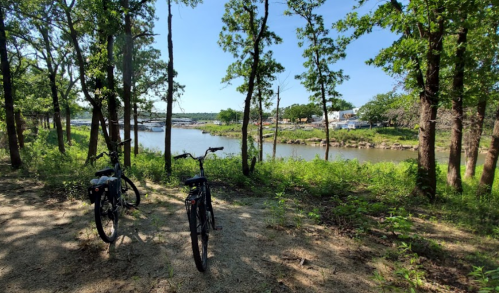 Two bicycles parked on a sandy path beside a river, surrounded by trees and greenery on a sunny day.