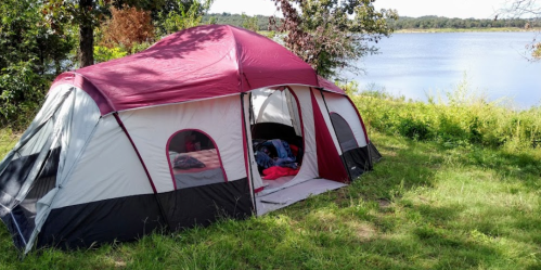A large red and gray tent set up on green grass by a calm lake, surrounded by trees and nature.