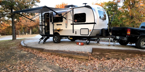 A white travel trailer parked on a gravel site surrounded by autumn leaves and trees. A black truck is attached.