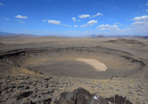 A vast, barren landscape featuring a large, circular crater surrounded by rocky terrain and distant mountains under a blue sky.