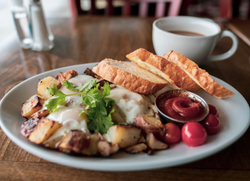 A plate of breakfast with eggs, potatoes, toast, cherry tomatoes, and a cup of coffee in the background.