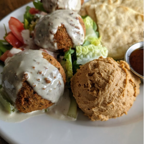 A plate featuring falafel balls drizzled with sauce, salad, pita bread, and a scoop of hummus.