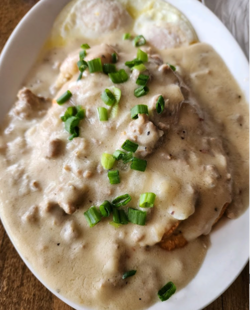 A plate of biscuits and gravy topped with chopped green onions, served with two fried eggs on the side.