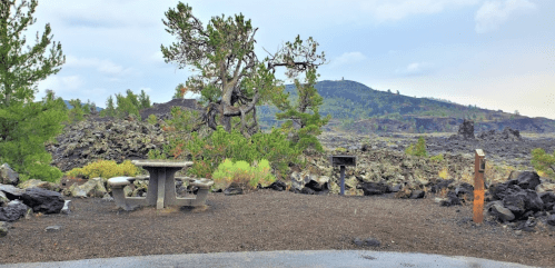 A picnic area surrounded by volcanic rock, with a table, benches, and a distant green hillside under a cloudy sky.