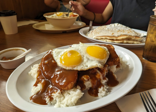 A plate of rice topped with fried chicken, brown gravy, and two sunny-side-up eggs, with pancakes in the background.