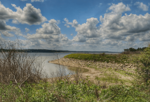 A serene lakeside view with a sandy shore, lush greenery, and a sky filled with fluffy clouds.