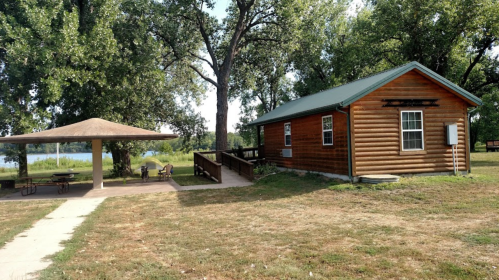 A wooden cabin beside a shaded picnic area, surrounded by trees and grass, near a body of water.