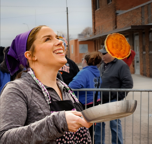 A woman in an apron and headscarf smiles as she flips a pancake in a frying pan at an outdoor event.