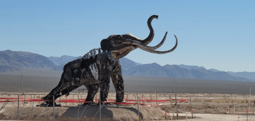 A large metal sculpture of a mammoth stands in a desert landscape with mountains in the background.