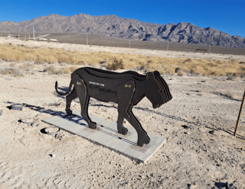 A metal sculpture of a lion walking in a desert landscape with mountains in the background.