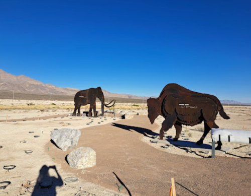 Metal sculptures of a mammoth and a bison in a desert landscape under a clear blue sky.