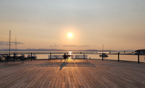 A serene sunset over a calm body of water, viewed from a wooden deck with boats in the distance.