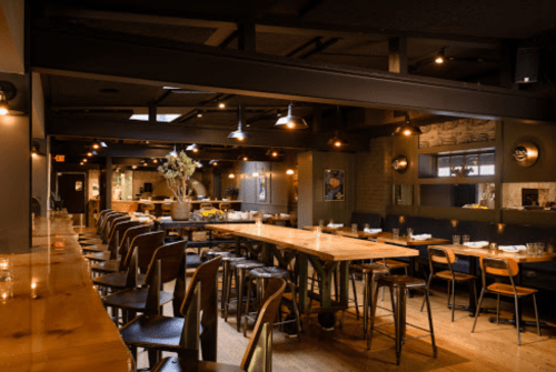 A modern restaurant interior featuring wooden tables, bar seating, and warm lighting.