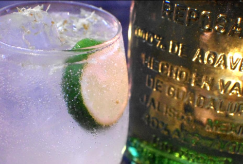 A close-up of a refreshing drink with lime slices and ice, next to a bottle of reposado tequila.