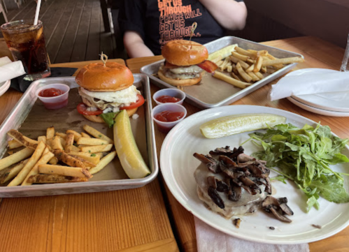 Two burgers with fries, a side of sautéed mushrooms, and a salad on a wooden table with drinks.