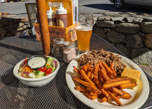 A plate of pulled pork, sweet potato fries, cornbread, and a salad, with a beer on a table outdoors.
