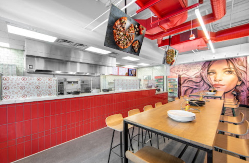 Modern restaurant interior with a red and white color scheme, featuring a long table, open kitchen, and vibrant wall art.