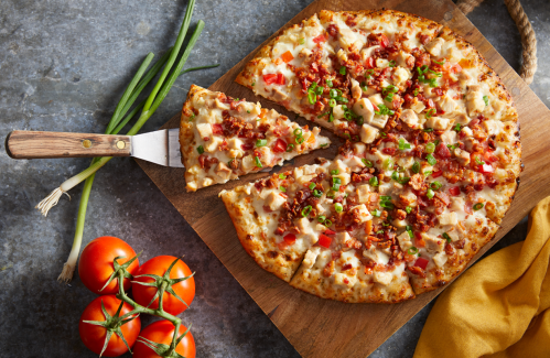A freshly baked pizza topped with bacon, chicken, tomatoes, and green onions, served on a wooden board with tomatoes nearby.