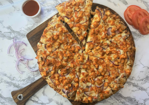 A round pizza topped with diced chicken, onions, and sauce, with a slice removed, on a wooden board. Sliced tomatoes and sauce nearby.