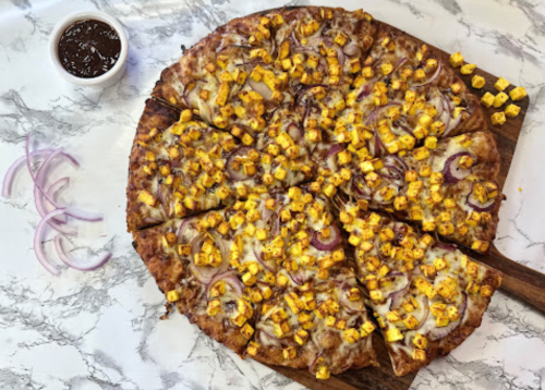 A pizza topped with pineapple, red onions, and cheese, sliced into pieces, with a small bowl of sauce on the side.