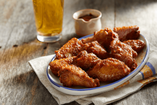 A plate of crispy chicken wings with a side of dipping sauce, accompanied by a glass of beer on a wooden table.
