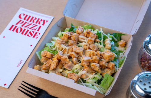 A salad with grilled chicken pieces in a takeout box, alongside a menu and condiments on a table.