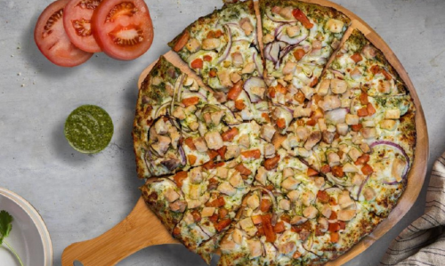 A sliced pizza topped with chicken, tomatoes, and onions, served with fresh tomato slices and green sauce on the side.