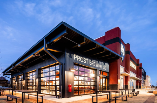 Exterior of Prost Brewing Co. with large windows, outdoor seating, and a colorful building at dusk.