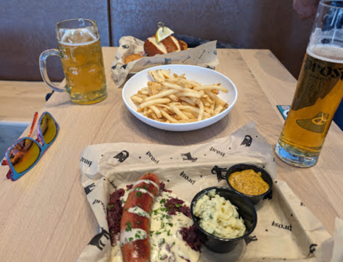 A table with a beer, fries, a hot dog with toppings, and a small bowl of mustard and sauce, plus colorful sunglasses.