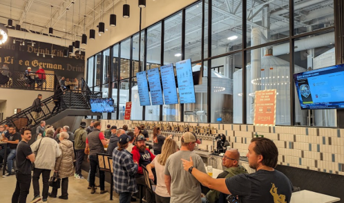 A busy brewery interior with a long bar, large screens displaying menus, and a crowd of patrons enjoying the atmosphere.