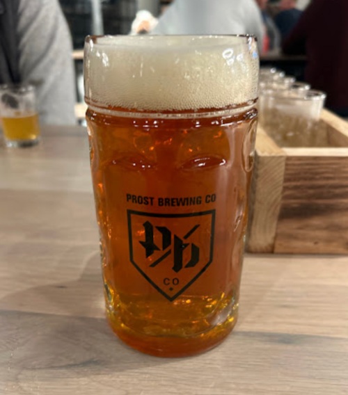 A glass of amber beer with a frothy head, featuring the Prost Brewing Co. logo, on a wooden table.