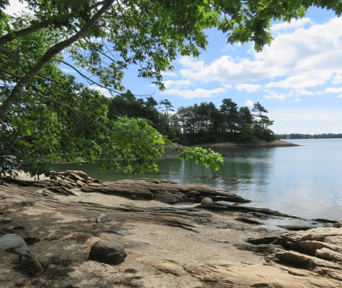 A serene lakeside view with rocky shorelines, trees, and a calm water surface under a partly cloudy sky.