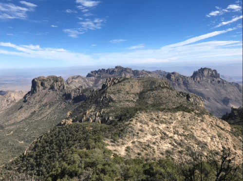 A panoramic view of rugged mountains and valleys under a clear blue sky with scattered clouds.