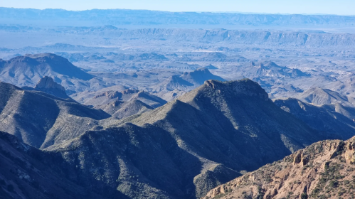 A panoramic view of rugged mountains and valleys under a clear blue sky, showcasing layered terrain and distant ridges.