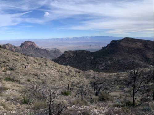 A scenic view of mountains and valleys under a blue sky, with hikers visible in the foreground.