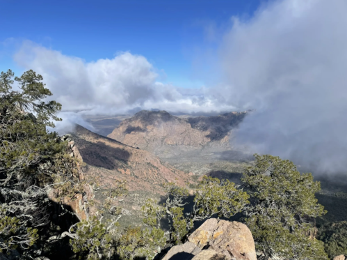 A panoramic view of a mountainous landscape with clouds and greenery in the foreground.