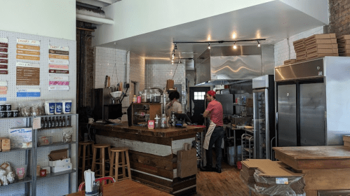 A cozy café interior with a wooden counter, two staff members working, and a menu displayed on the wall.