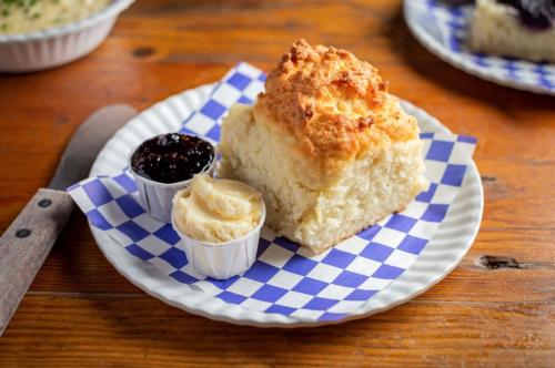 A piece of fluffy biscuit on a plate with butter and jam in small cups, set on a checkered paper.