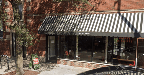 Exterior of a coffee shop named "Bang Bang," featuring a striped awning and large windows, surrounded by trees.