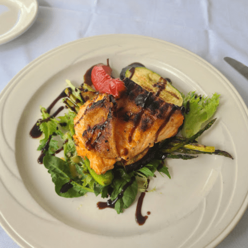 Grilled salmon on a bed of mixed greens, garnished with asparagus, zucchini, and a drizzle of balsamic reduction.