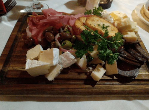 A wooden platter featuring assorted cheeses, cured meats, olives, and garnished with parsley and toasted bread.