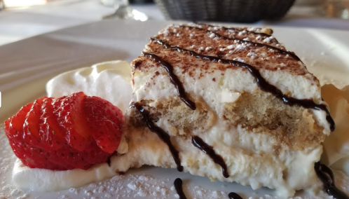 A slice of tiramisu topped with cocoa powder, drizzled with chocolate, and served with a strawberry and whipped cream.